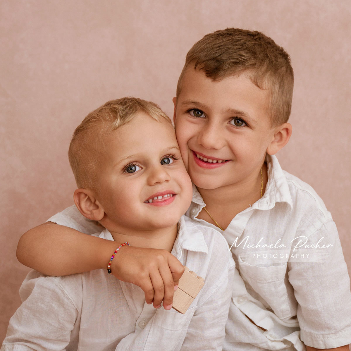 Zwei Brüder sitzen eng beieinander, der ältere umarmt den jüngeren liebevoll, beide lachen fröhlich in die Kamera.